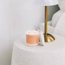 Candle with a peach label on a textured surface next to a gold lamp.