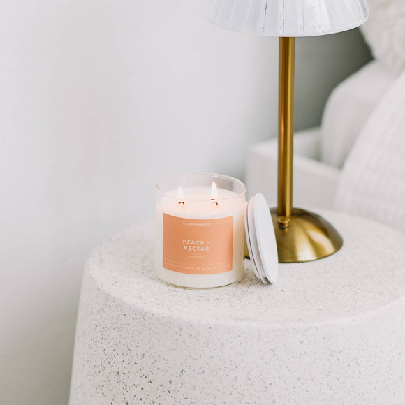 Candle with a peach label on a textured surface next to a gold lamp.
