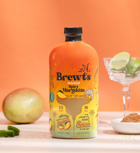 Bottle of Brewt's Mango Spicy Margarita with ingredients on a table against an orange background