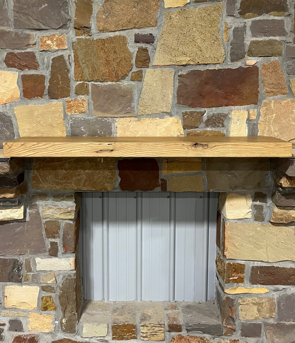 A beam style fireplace mantel, made from white oak wood, mounted on a stone wall.