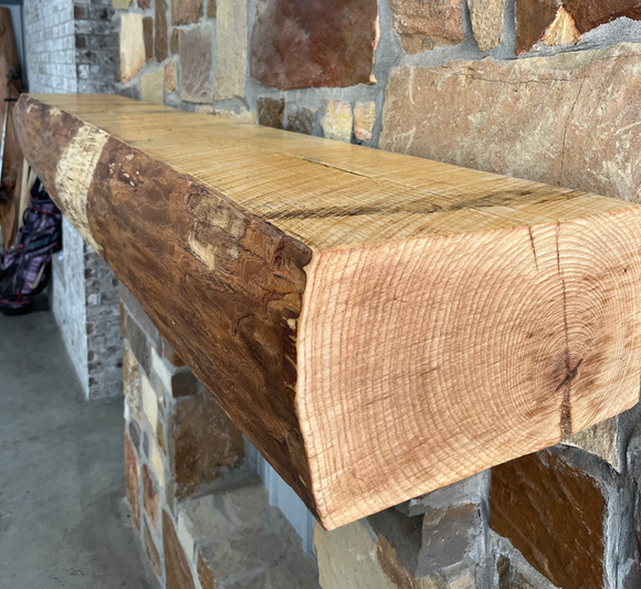 A live edge style fireplace mantel, made from ash wood, mounted on a stone wall.