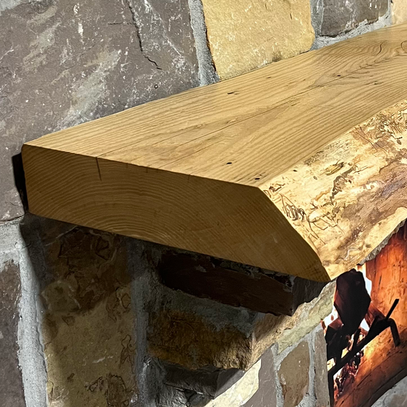 A live edge style fireplace mantel, made from ash wood, mounted on a stone wall.