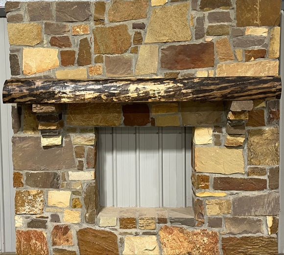 A live edge style fireplace mantel, made from walnut wood, mounted to a rock wall.
