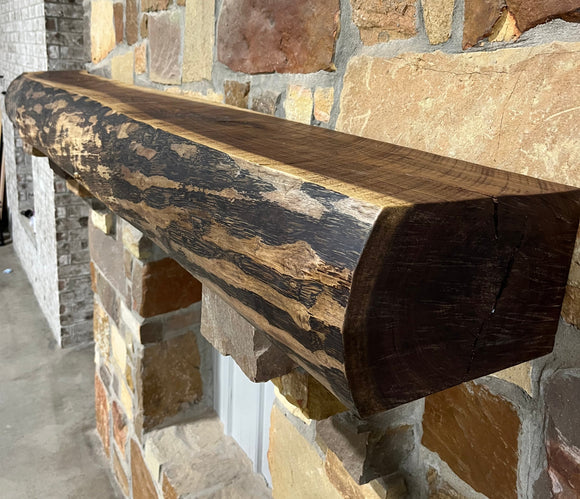 A live edge style fireplace mantel, made from walnut wood, mounted to a rock wall.