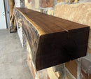 A live edge style fireplace mantel, made from walnut wood, mounted to a rock wall.