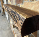 A live edge style fireplace mantel, made from walnut wood, mounted to a rock wall.