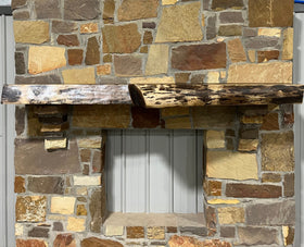 A live edge style fireplace mantel, made from walnut wood, mounted to a rock wall.