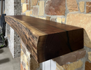 A live edge style fireplace mantel, made from walnut wood, mounted to a rock wall.