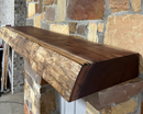 A live edge style fireplace mantel, made from walnut wood, mounted to a rock wall.