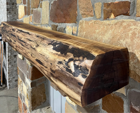 A live edge style fireplace mantel, made from walnut wood, mounted to a rock wall.