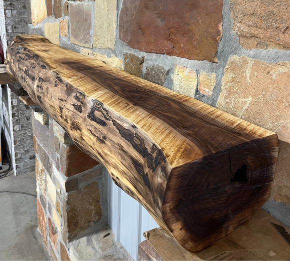 A live edge style fireplace mantel, made from walnut wood, mounted to a rock wall.