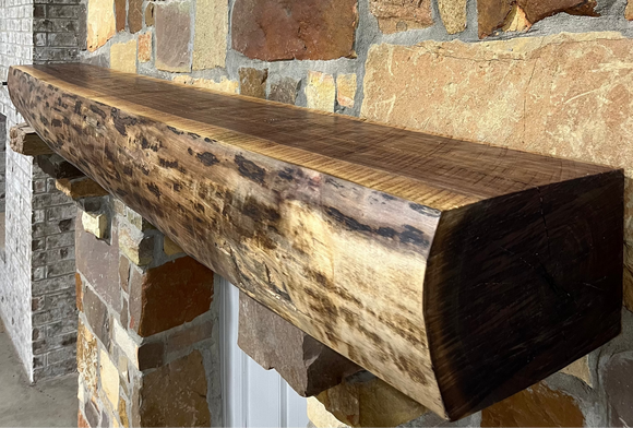 A live edge style fireplace mantel, made from walnut wood, mounted to a rock wall.