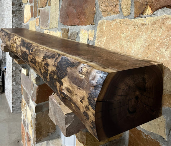 A live edge style fireplace mantel, made from walnut wood, mounted to a rock wall.