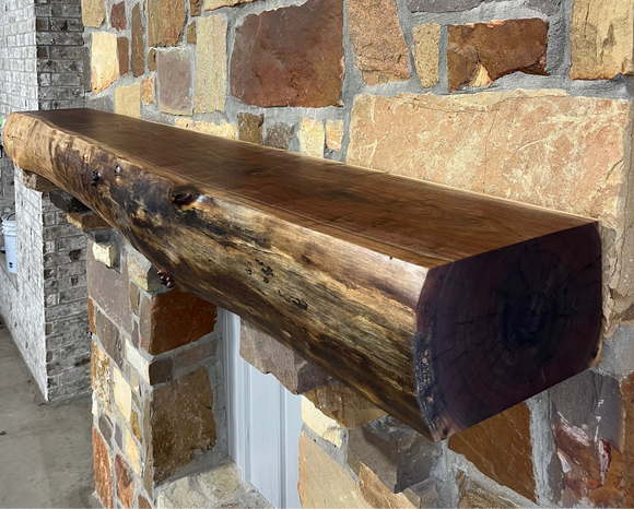 A live edge style fireplace mantel, made from walnut wood, mounted to a rock wall.