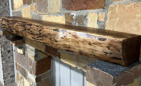 A live edge style fireplace mantel, made from walnut wood, mounted to a rock wall.