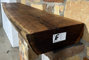 A live edge style fireplace mantel, made from walnut wood, mounted to a rock wall.