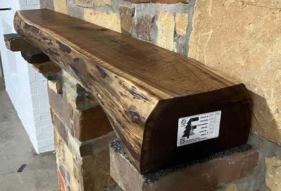 A live edge style fireplace mantel, made from walnut wood, mounted to a rock wall.