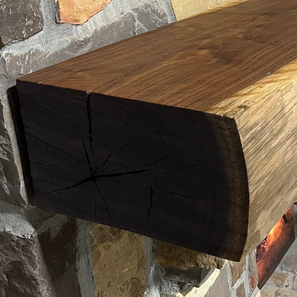 A live edge style fireplace mantel, made from walnut wood, mounted to a rock wall.