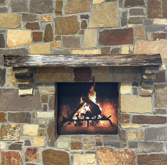 A live edge style fireplace mantel, made from walnut wood, mounted to a rock wall.