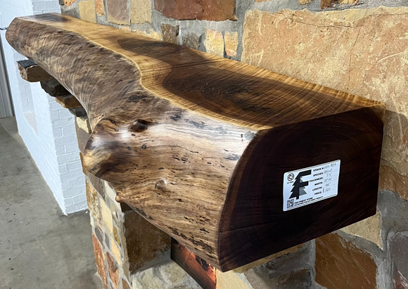 A live edge style fireplace mantel, made from walnut wood, mounted to a rock wall.