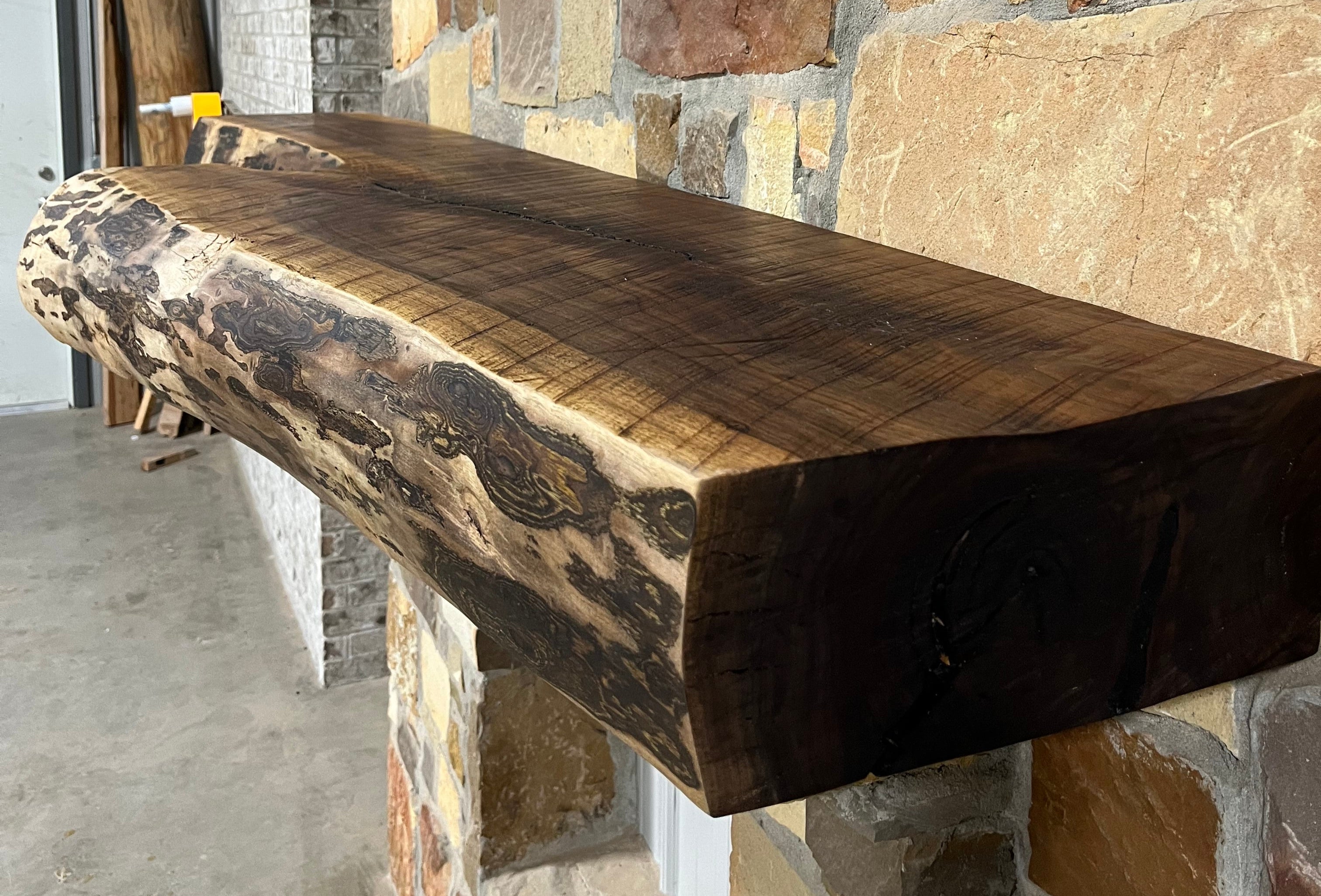 A solid walnut wood fireplace mantel with the side of the tree exposed on a stone wall.