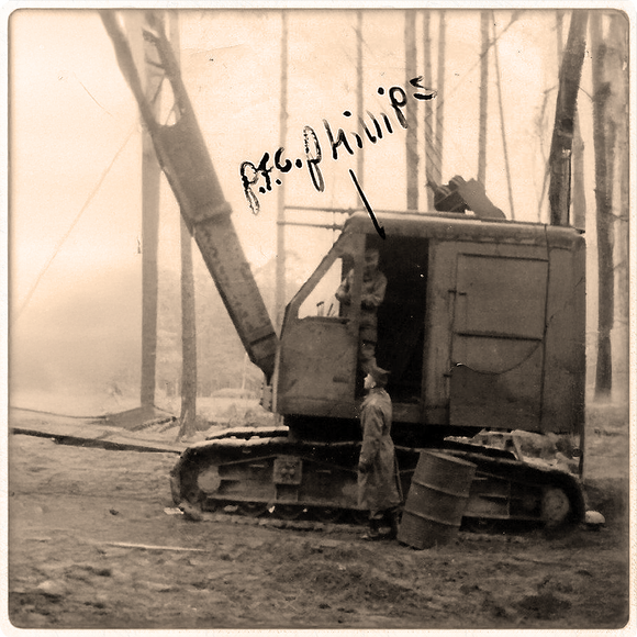 Vintage German logging crane black and white photo of a large piece of industrial equipment with a person standing nearby, set against a forest background.